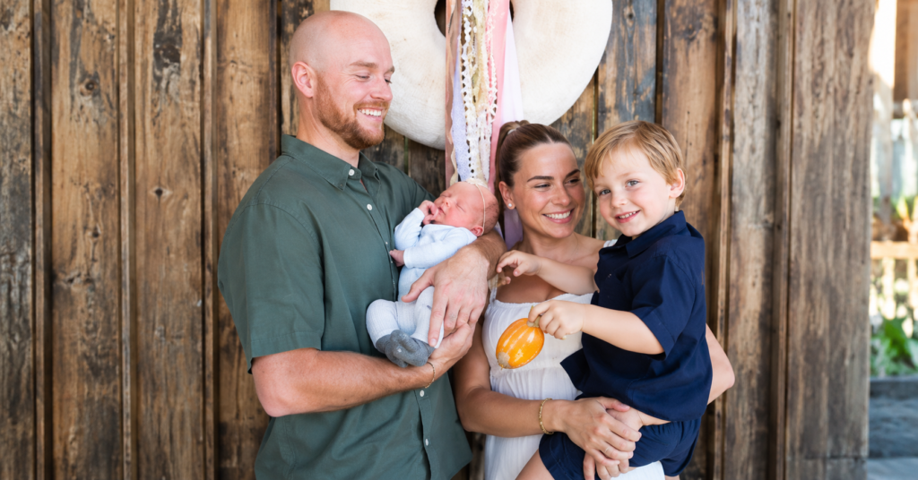 Trigger Kiser family portrait featuring a happy family of four posing together with their newborn baby and young child in a warm rustic setting.