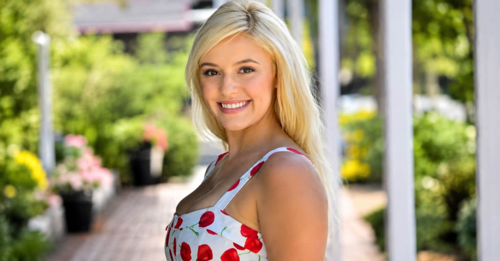 Portrait of Kylie Page smiling outdoors in a white floral summer dress, standing in a sunlit garden walkway with a softly blurred green background.