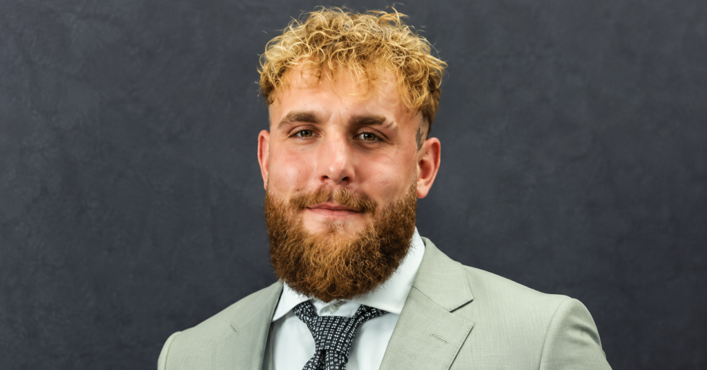 Close-up portrait of Jake Paul with curly blond hair and a full beard, wearing a light gray suit, white shirt, and patterned tie against a blue backdrop.