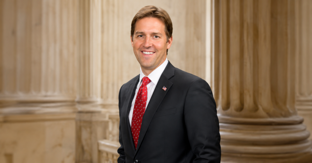 Official portrait of Ben Sasse smiling in a dark suit, white shirt, and red tie with marble columns in the background.