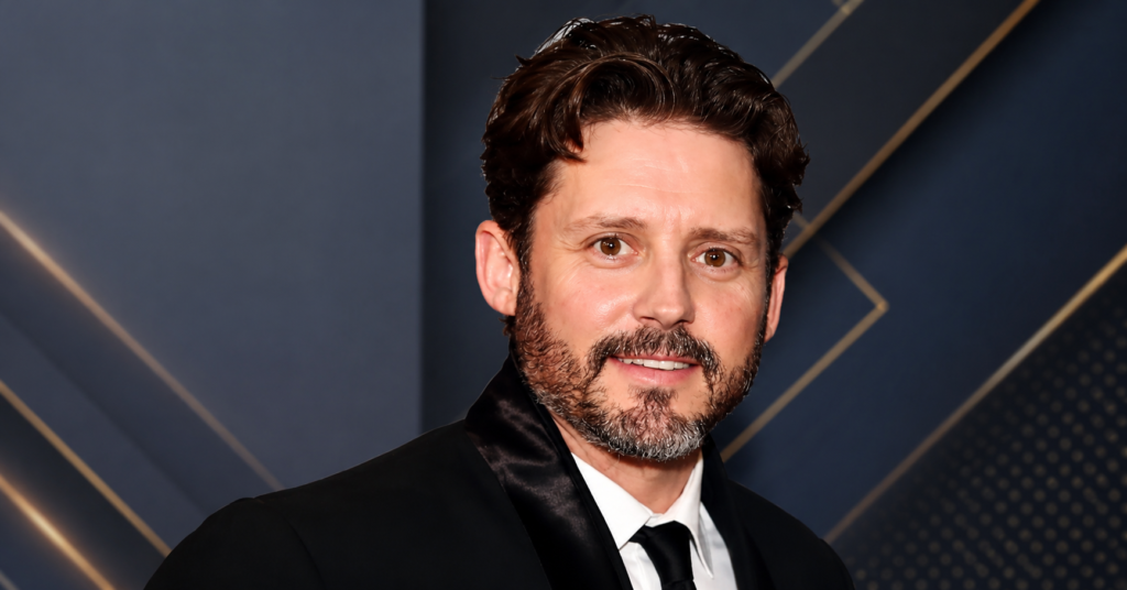 Brandon Blackstock wearing a black tuxedo at a red carpet event with a gray background.